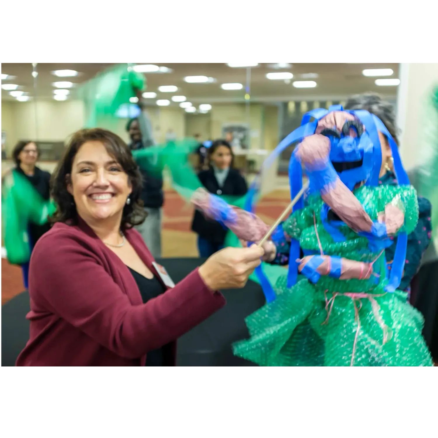 A woman looks over her right shoulder at the camera, smiling. Her upper body is visible, right arm bent: she holds a thin dowel with a streamer attached. A costumed figure across from her appears to also be waving brightly colored streamers; they may be part of its costume. Colorful streamers swirl behind them.