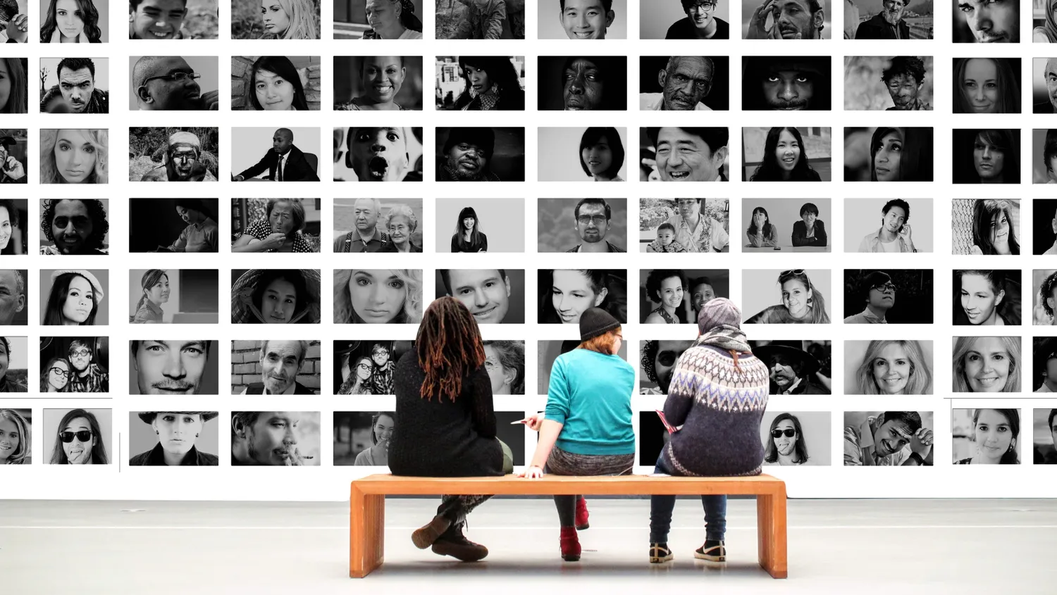 Three people sit on a bench viewing a large wall display of diverse portraits, representing community, diversity, and human connection.