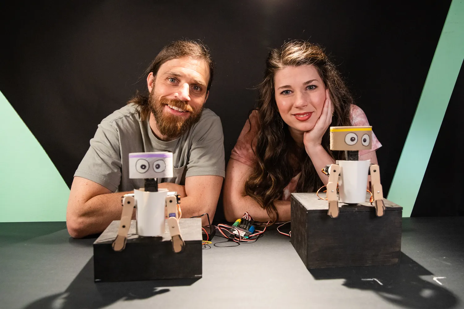 Matt Chilbert, left, and Kelsey Derringer, right, face the camera. They are both white adults with dark hair. in front of them are two small robot figures.