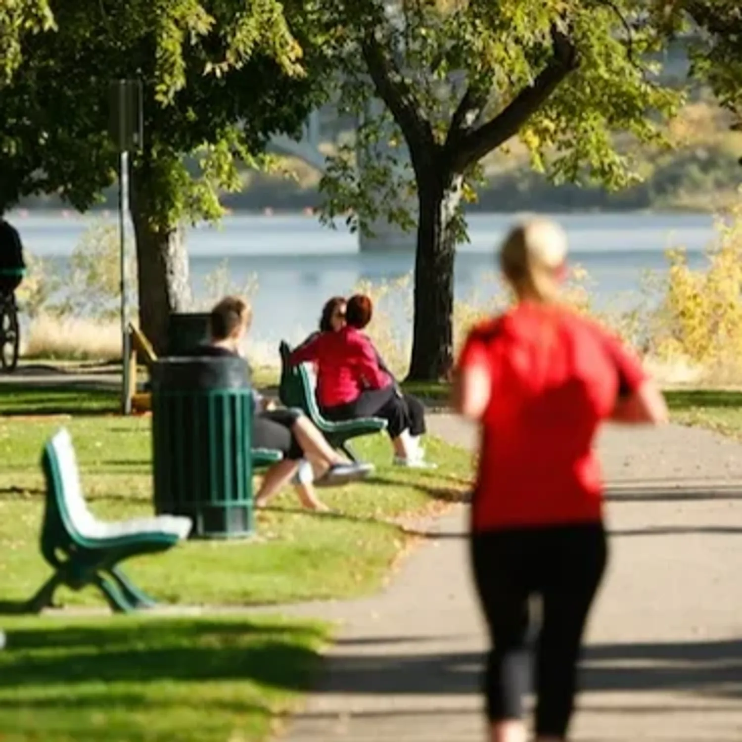 People walk, run, and cycle along a tree-lined riverside trail beside the South Saskatchewan River.