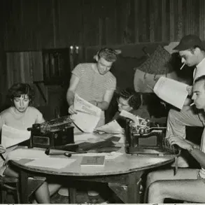 Vintage black & white photo of a group of informally dressed people (men and women) around a small table. Those standing are holding sheets of paper; those seated are typing.