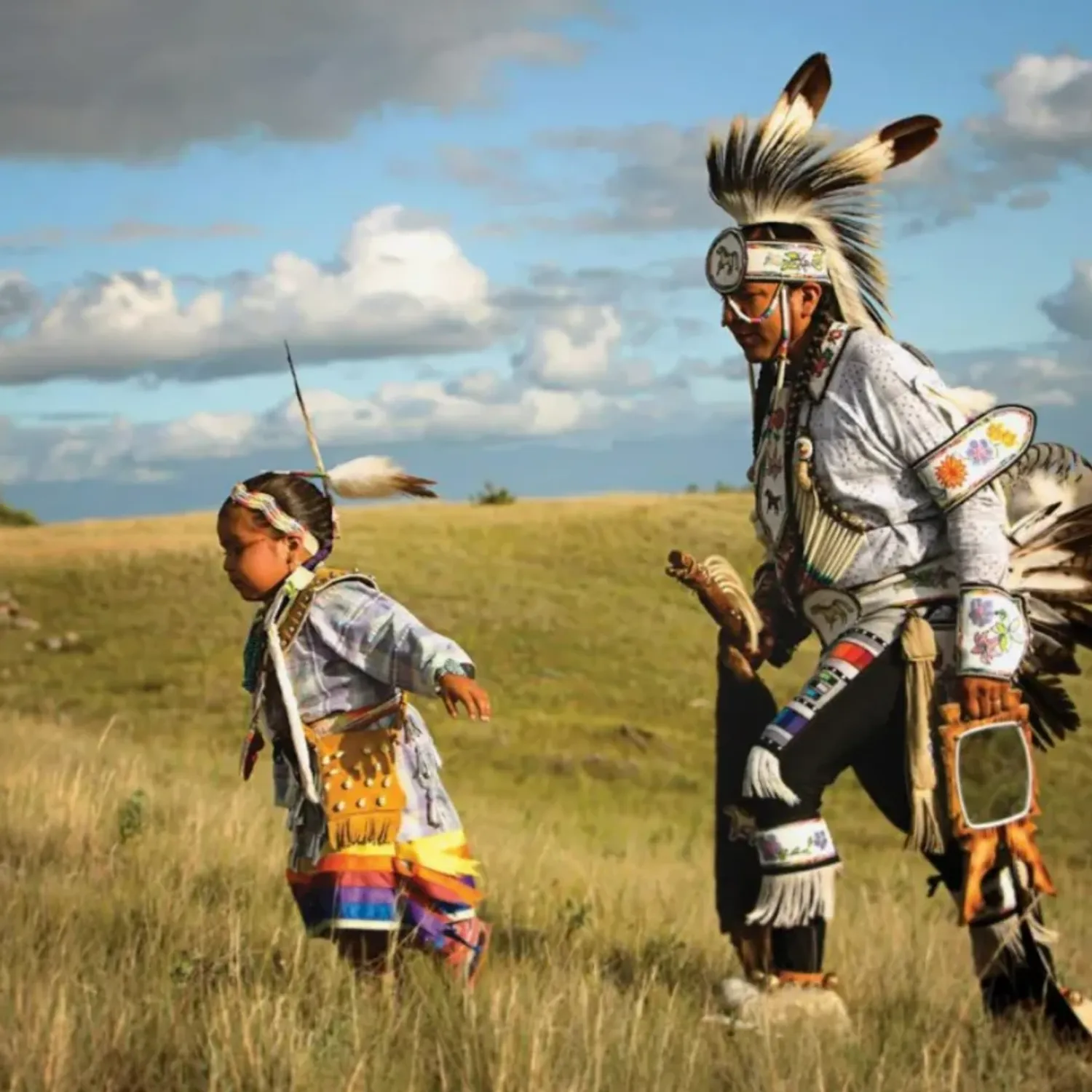 An Indigenous adult and child wearing traditional regalia walk together across a grassy prairie landscape under a dramatic sky.