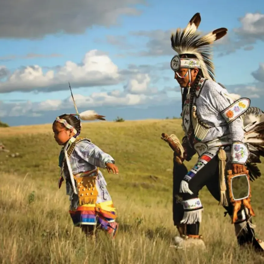 An Indigenous adult and child wearing traditional regalia walk together across a grassy prairie landscape under a dramatic sky.