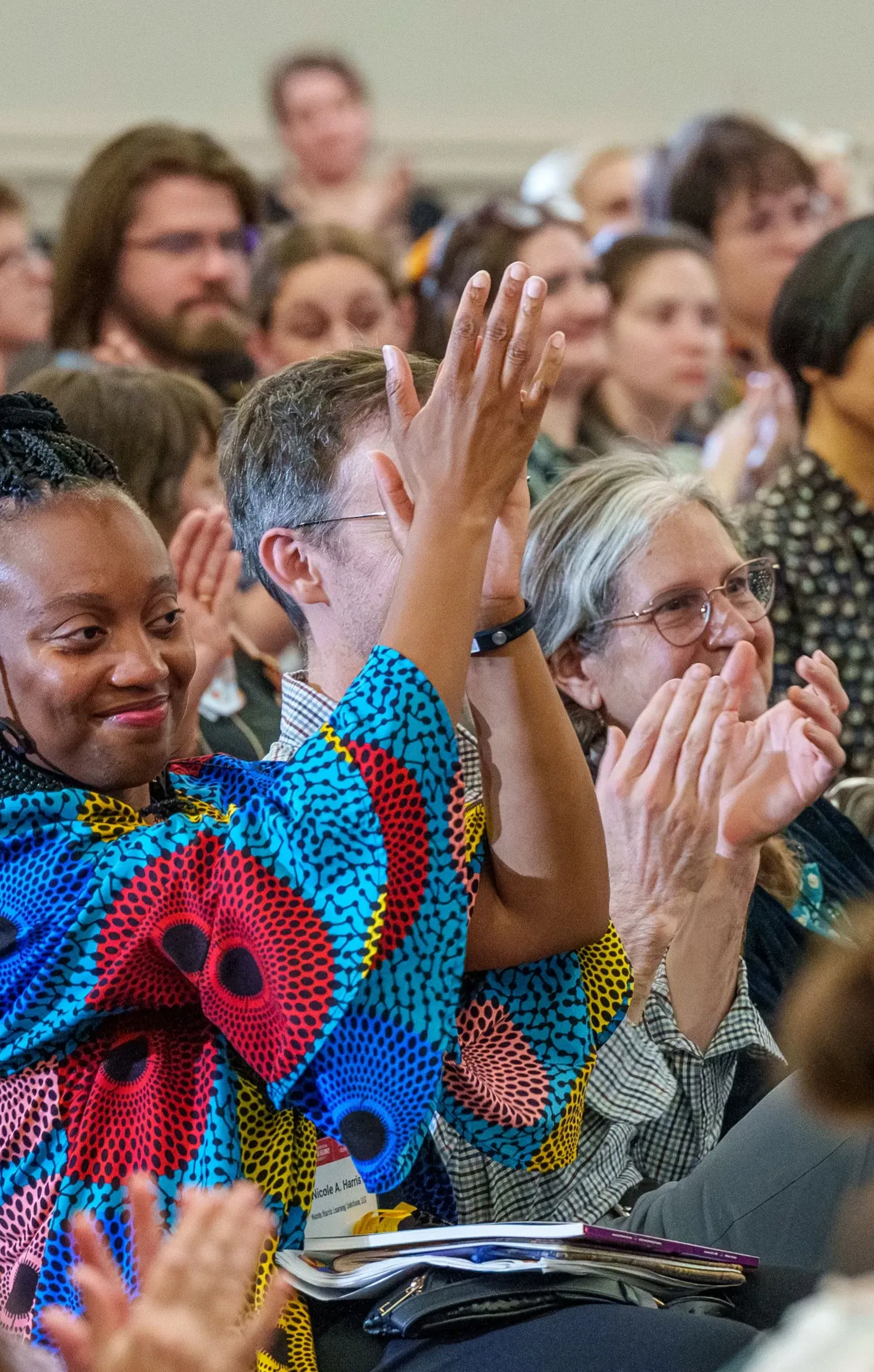 Photo of a smiling crowd at a lecture. A woman in colorful clothing raises her hands in applause. Photo by Josh Weilepp, for the American Folklore Society.