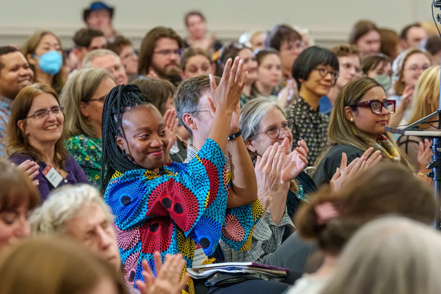Photo of a smiling crowd at a lecture. A woman in colorful clothing raises her hands in applause. Photo by Josh Weilepp, for the American Folklore Society.
