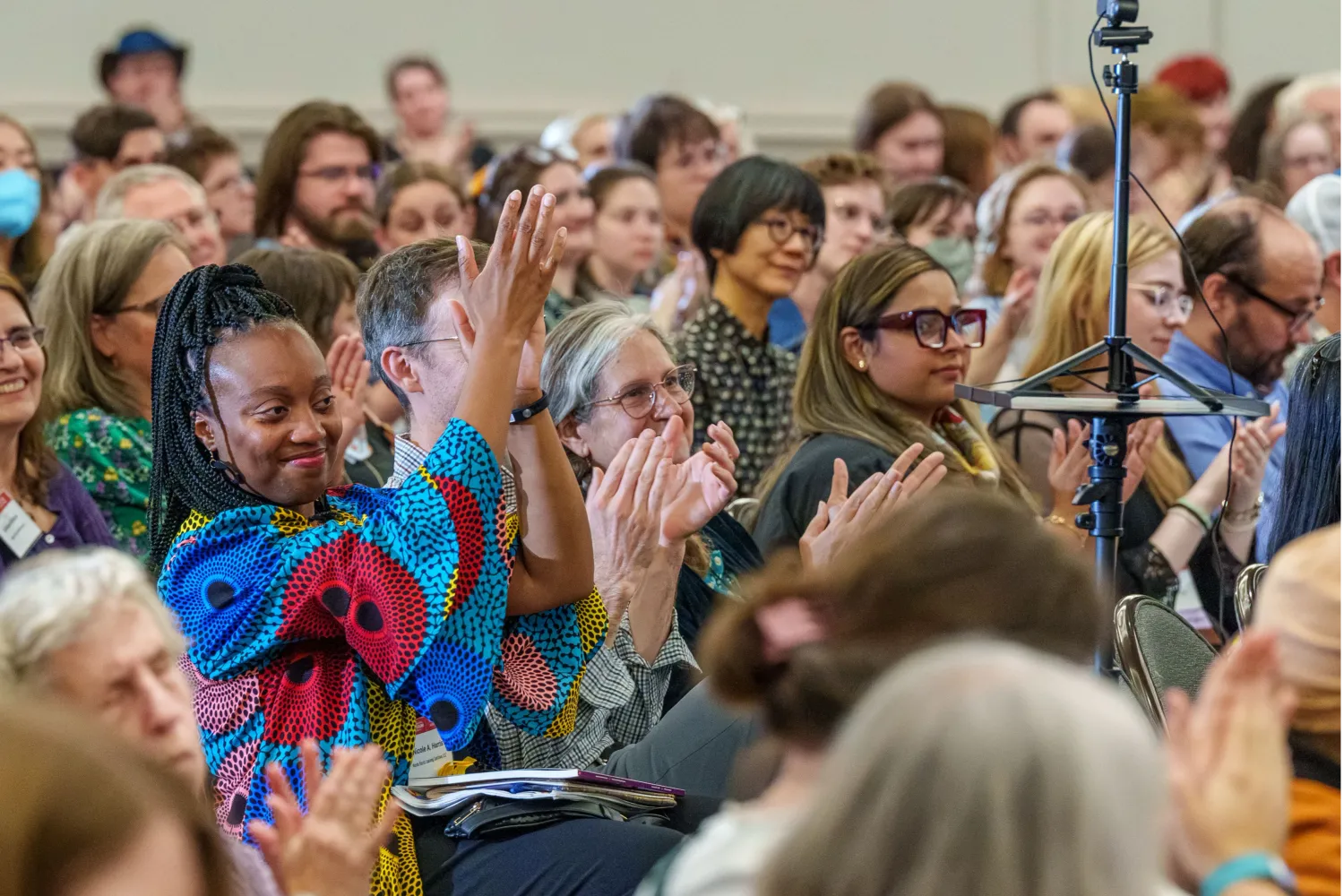 Photo of a smiling crowd at a lecture. A woman in colorful clothing raises her hands in applause. Photo by Josh Weilepp, for the American Folklore Society.
