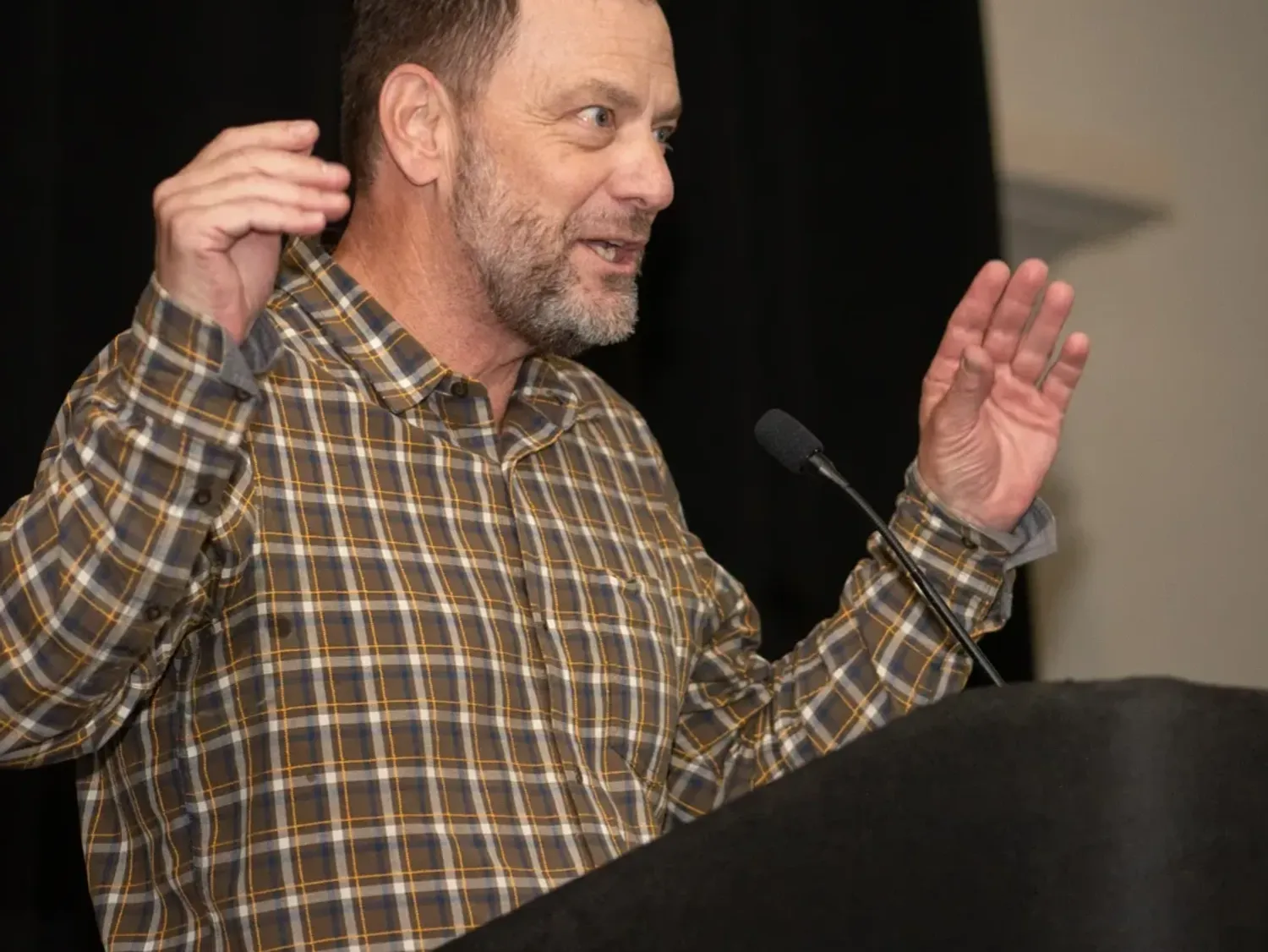A bearded man in a western plaid shirt stands behind a lectern, gesturing with both arms.
