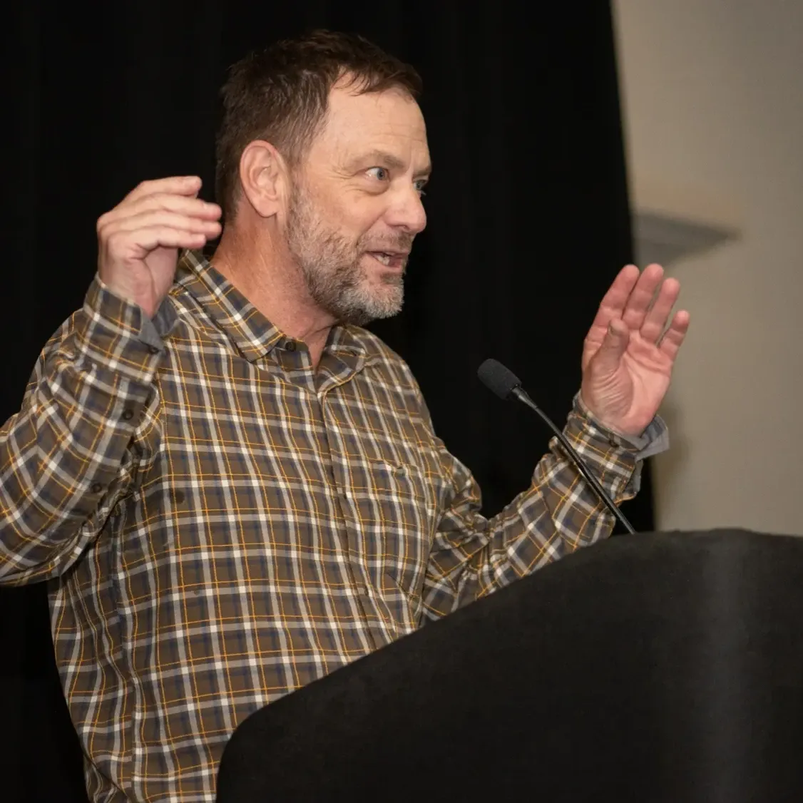 A bearded man in a western plaid shirt stands behind a lectern, gesturing with both arms.