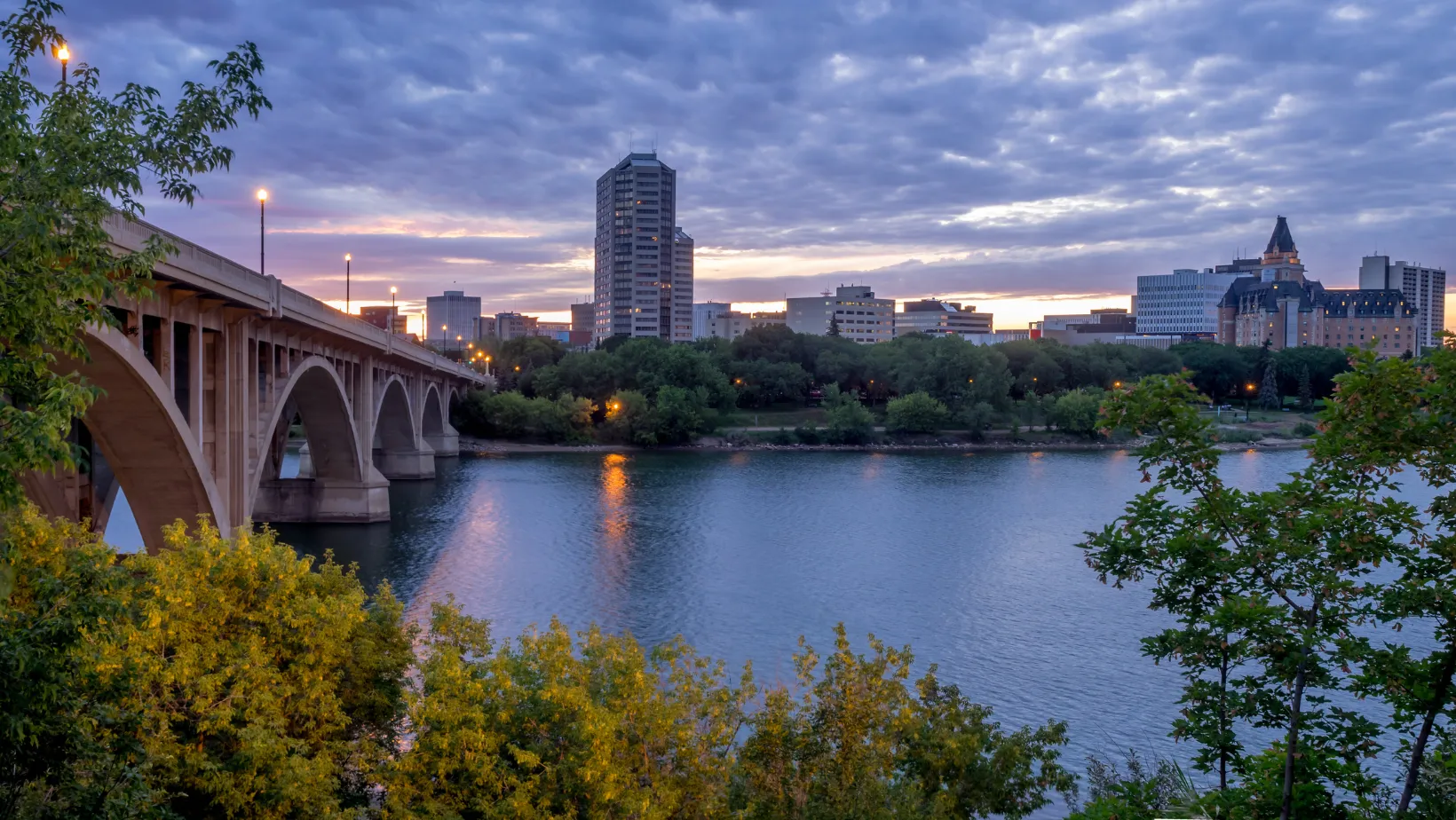 View of Saskatoon at dusk, featuring a concrete bridge crossing the South Saskatchewan River, trees along the riverbank, and the downtown skyline beneath a cloudy evening sky.
