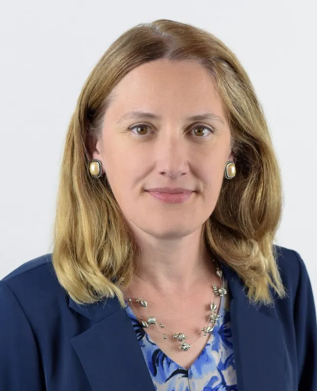 Professional headshot of Monica Sarghie looking at the camera with a gentle smile, wearing a navy blue blazer over a blue patterned blouse, pearl-style earrings, and a silver necklace. She has shoulder-length blonde hair and is photographed against a plain white background.