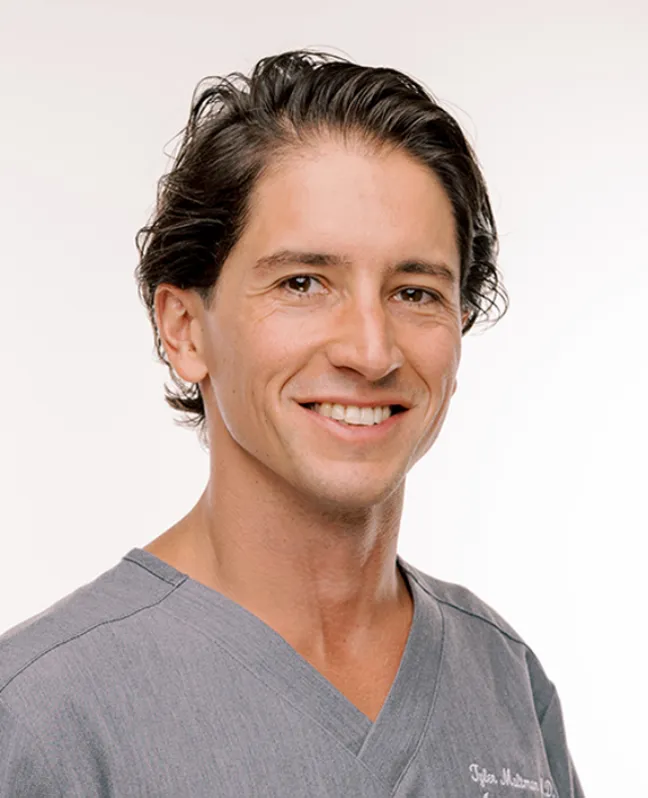 Headshot of Dr. Tyler Maltman smiling at the camera, wearing grey medical scrubs against a white background.