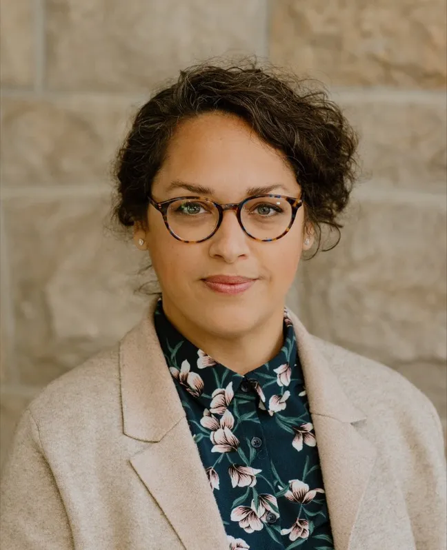Professional headshot of Dr. Meera Kachroo smiling gently at the camera, wearing patterned glasses, a dark floral blouse, and a beige blazer. She has short, curly dark hair and stands against a neutral stone wall background.