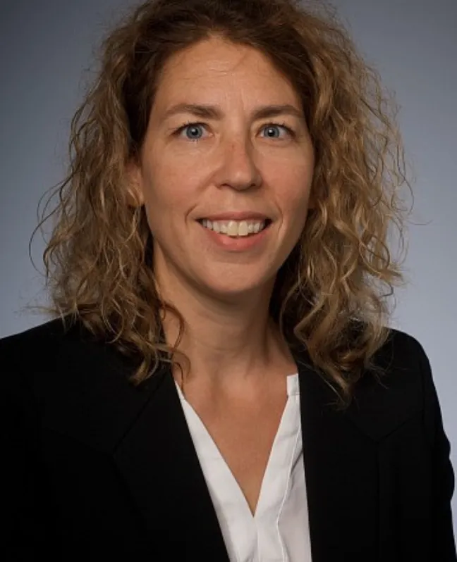 Professional headshot of Chantal Morin smiling at the camera, wearing a black blazer over a white blouse. She has shoulder-length, curly light brown hair and is photographed against a neutral grey background.