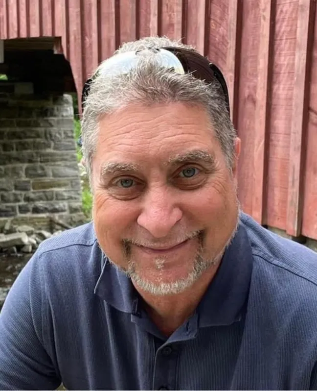 Photograph of Dr. Pierre Haddad smiling at the camera, wearing a navy blue polo shirt with sunglasses resting on his head. He has short grey hair and a neatly trimmed grey beard and is seated outdoors in front of a rustic red wooden building and stone wall.