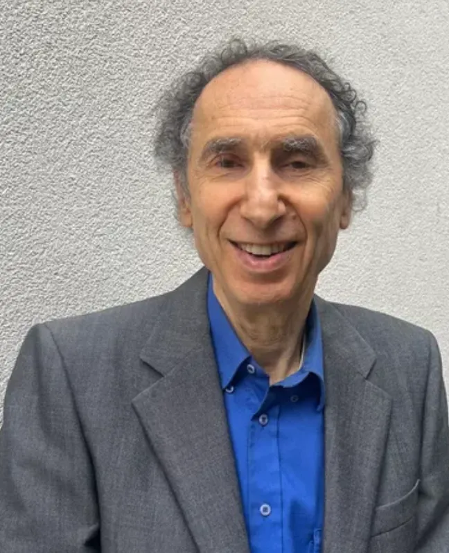 Portrait of Dr. Michael Epstein smiling at the camera, wearing a grey blazer over a bright blue shirt and standing against a light-coloured wall.